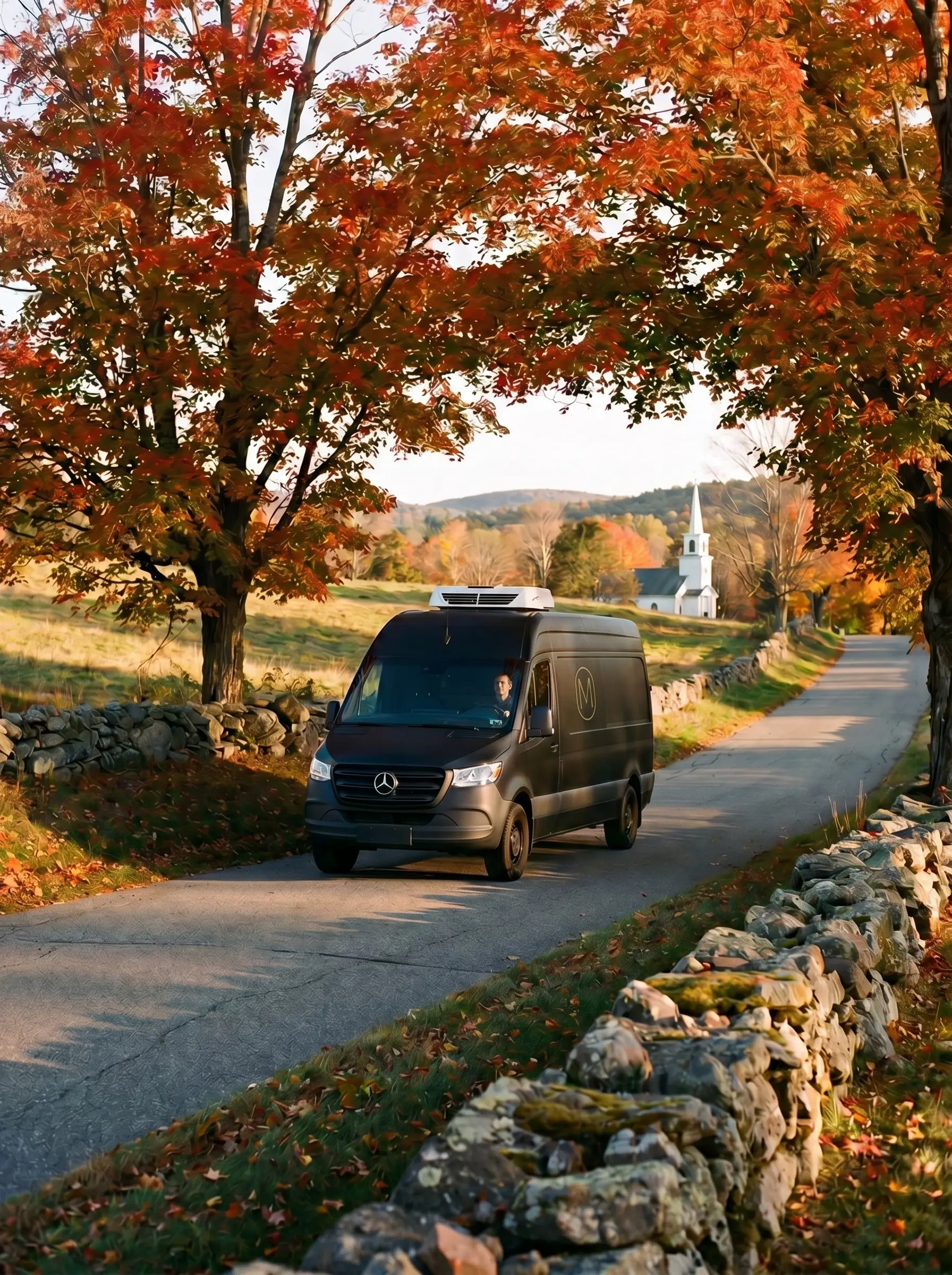 Morabeza matte-black Sprinter delivery van with a small gold M-in-capsule emblem on the side and a roof-mounted refrigeration unit, driver visible behind the wheel, traveling a winding central Massachusetts country road lined with stone walls and sugar maples in autumn foliage at golden hour