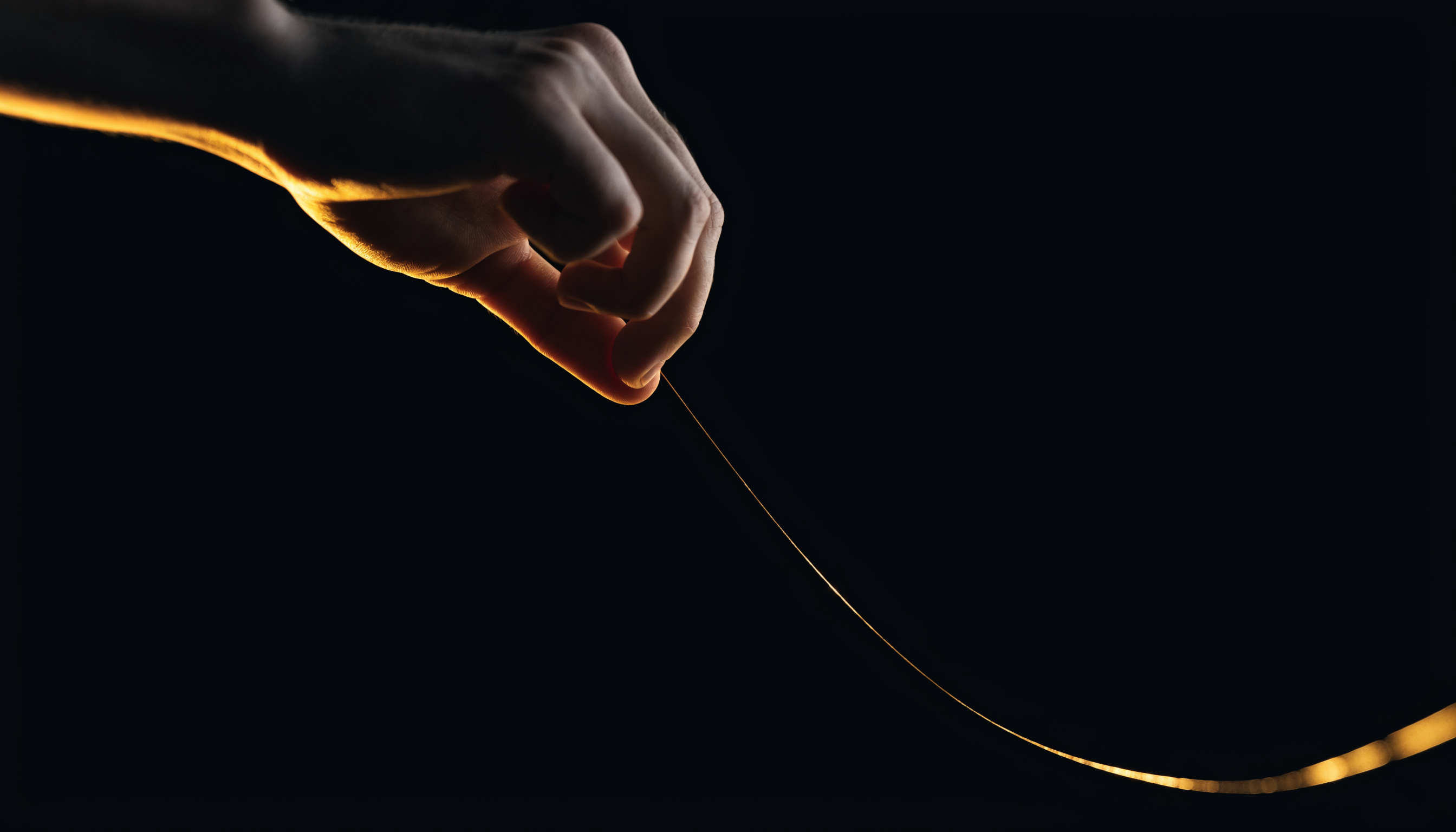 Uma mão soltando um fio dourado que estava esticado contra um fundo preto, o fio ficando frouxo no ar sob luz cinematográfica quente, metáfora de tirar a pressão no fechamento da oferta