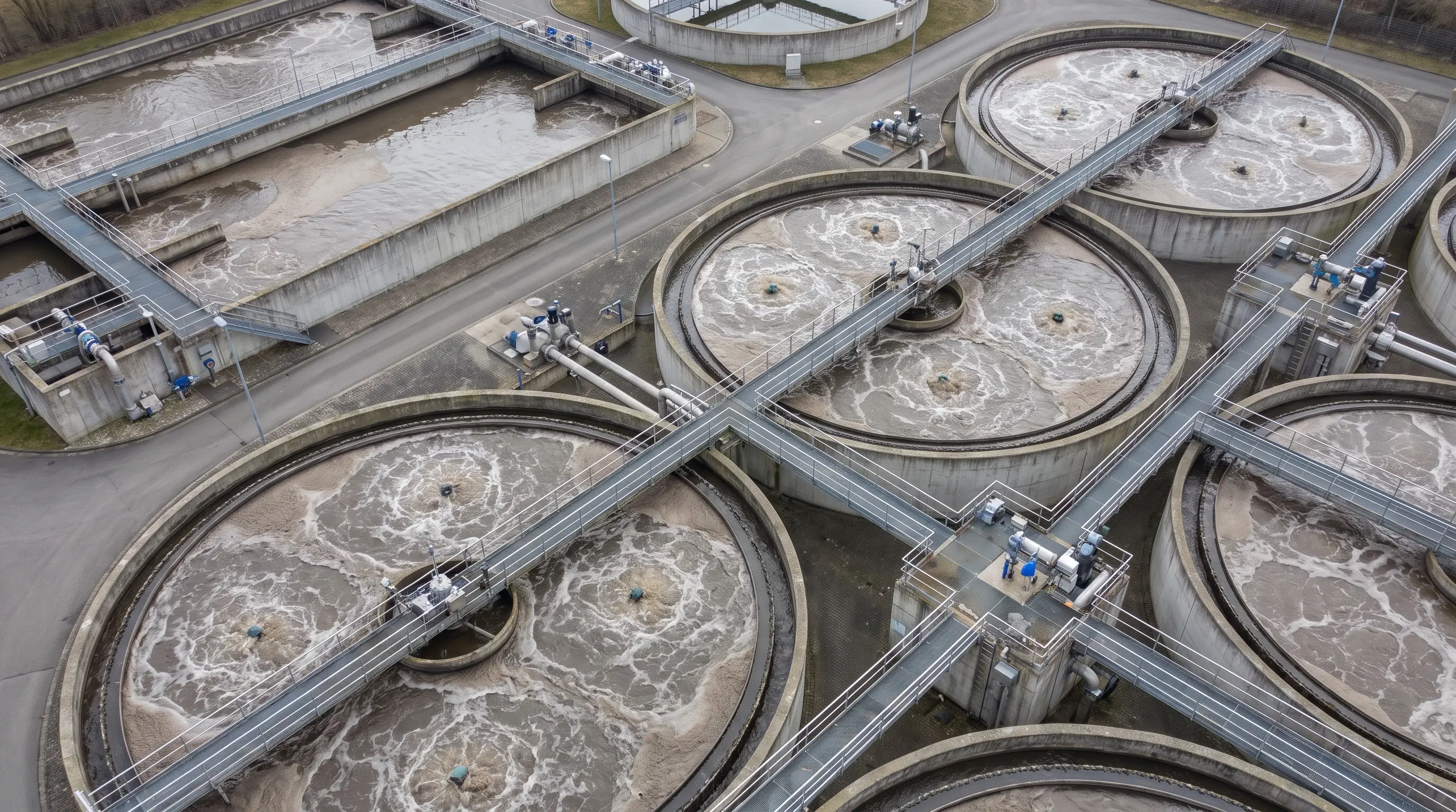 Luftbild einer kommunalen Kläranlage mit runden und rechteckigen Belebungsbecken, aufgewühltem Wasser und Schaum