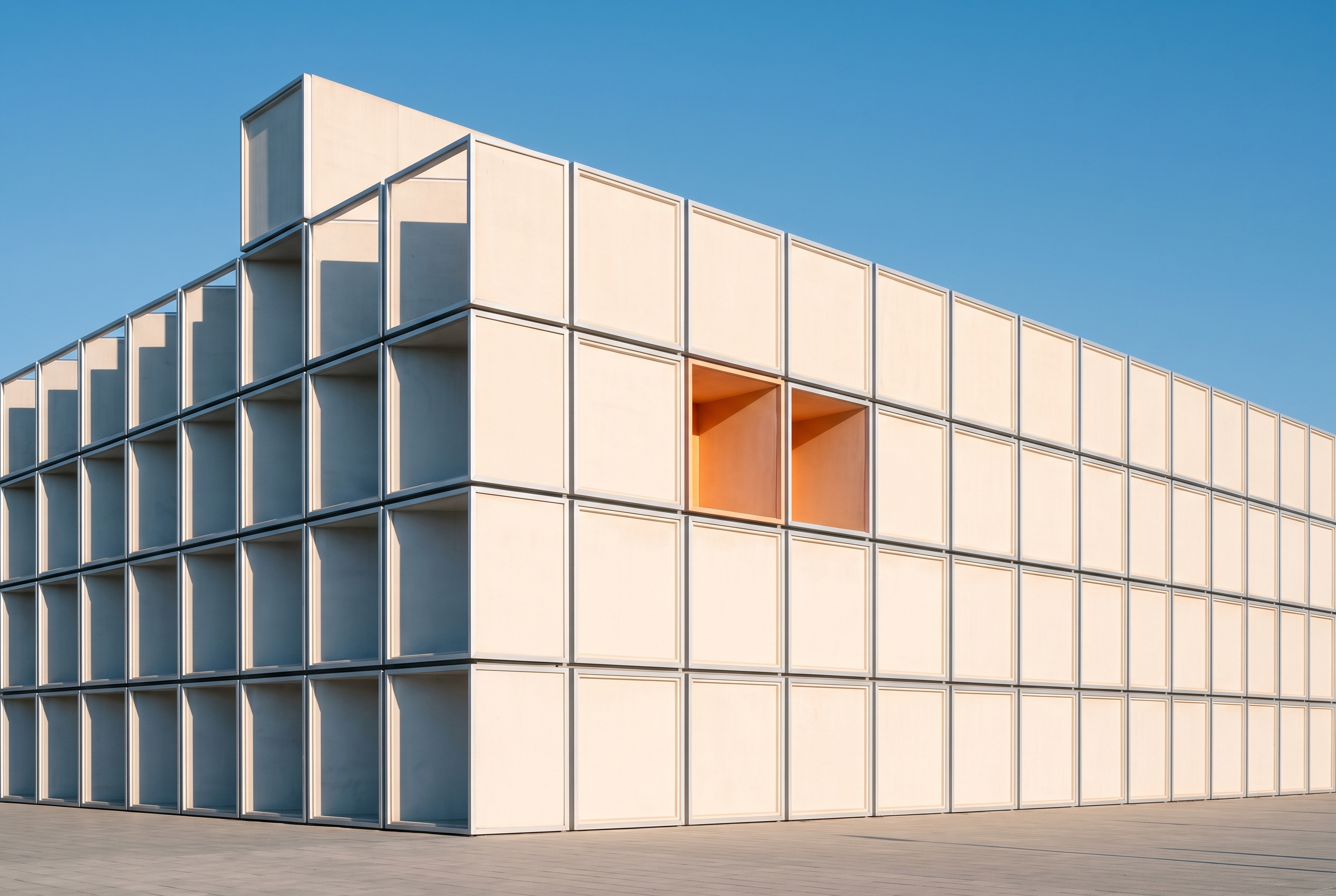 A precisely ordered facade of stacked pale concrete volumes with one module lit from within