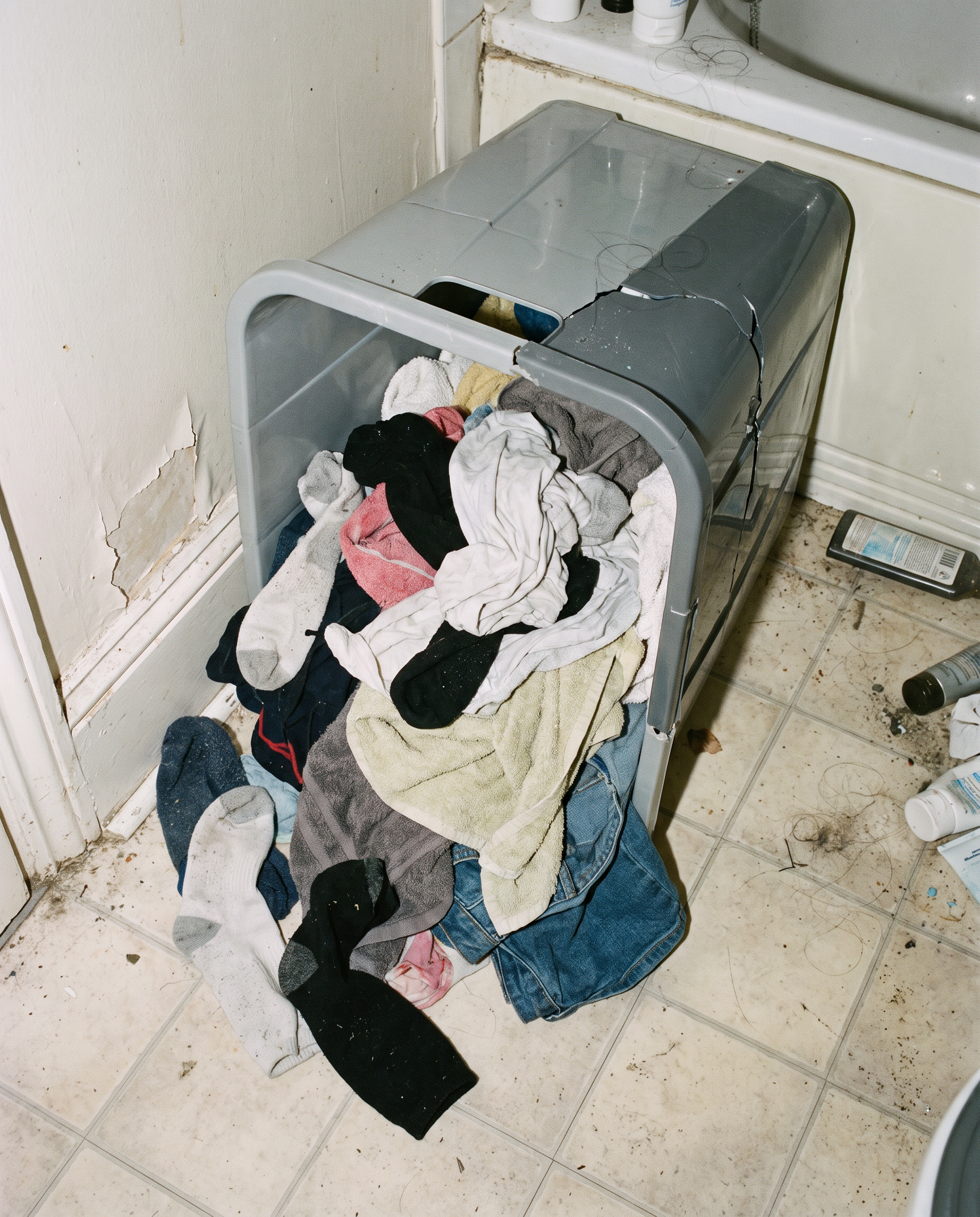 A cracked plastic laundry bin overflowing with clothes in a cluttered corner