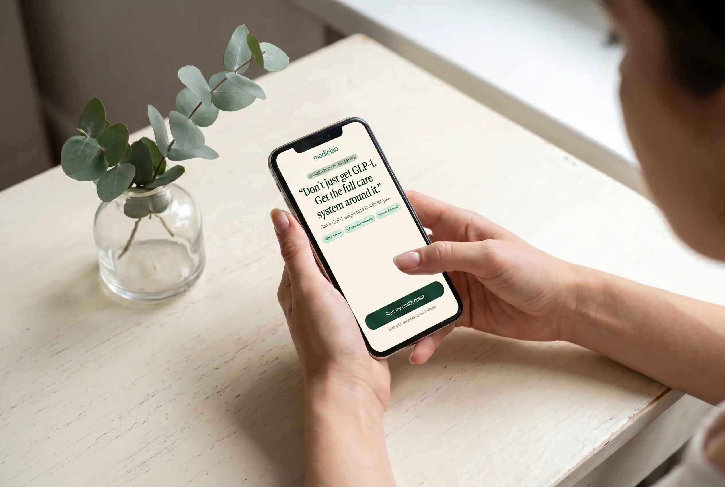 A patient holding an iPhone showing the MedicLab GLP-1 health check intake screen