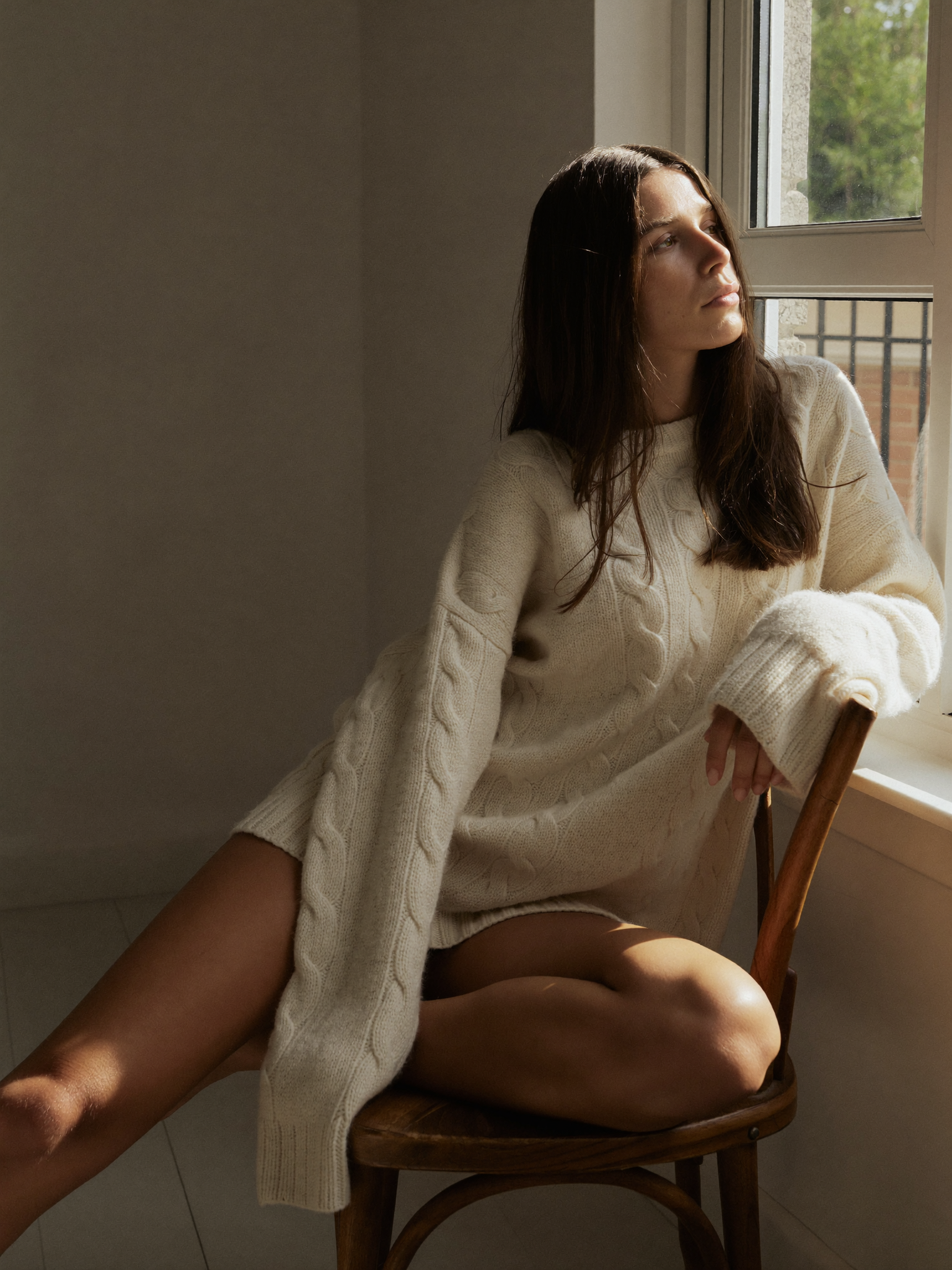 Woman in an oversized cream knit sweater by a sunlit window