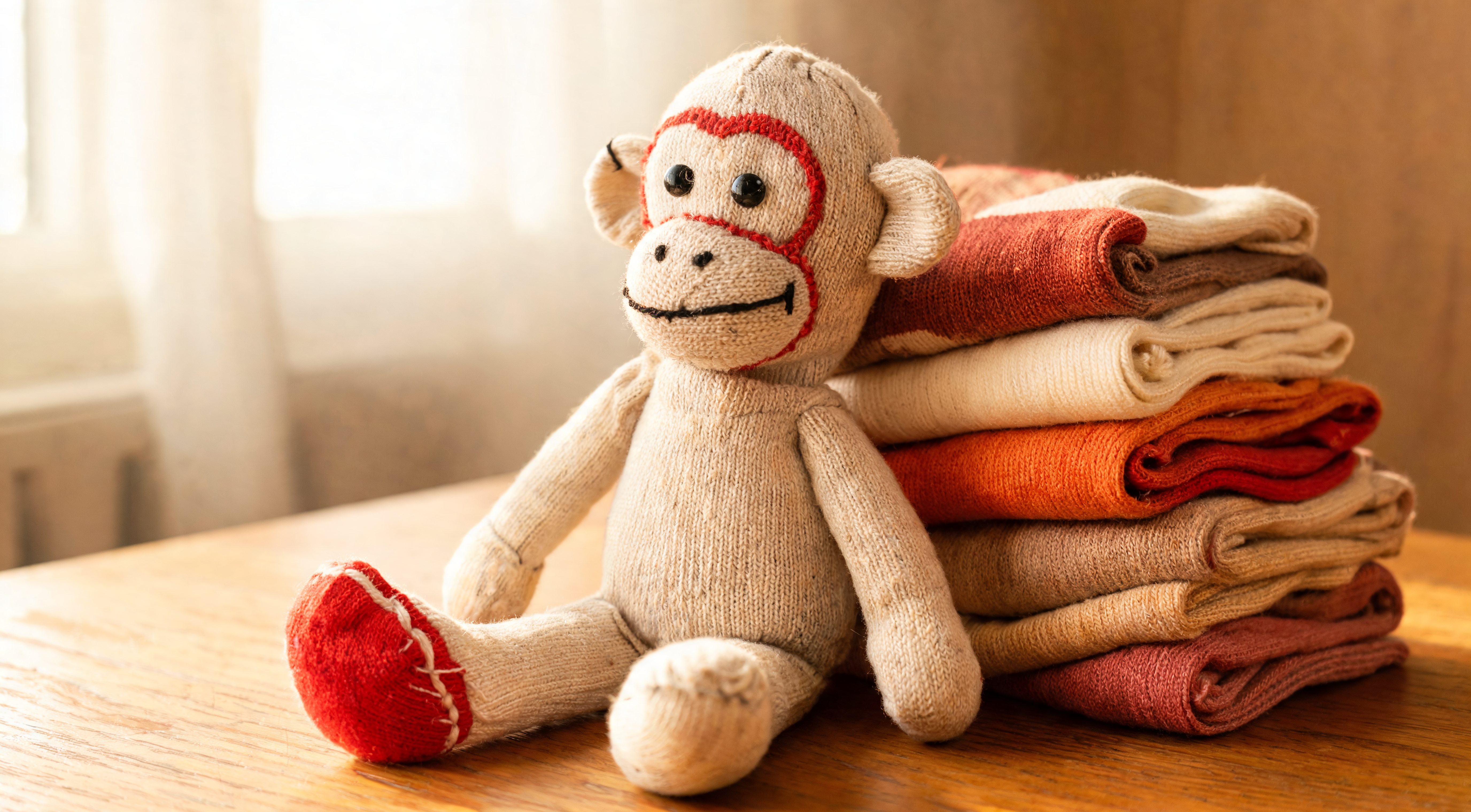A hand-stitched sock monkey doll with a red heel patch, propped against a stack of folded colorful socks on a wooden table