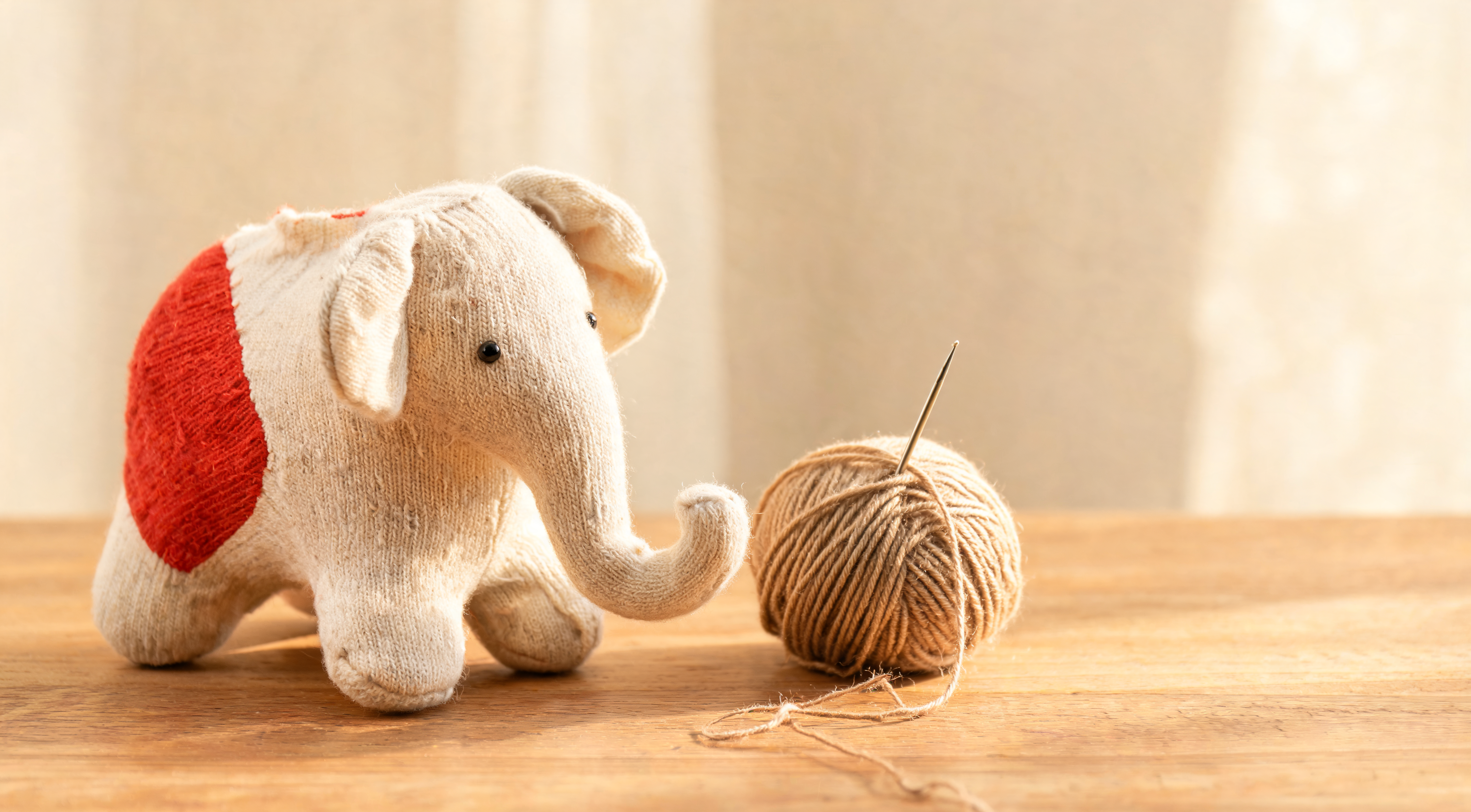 A handmade sock elephant toy sewn from a red heel sock, sitting on a wooden table next to a ball of yarn and a needle