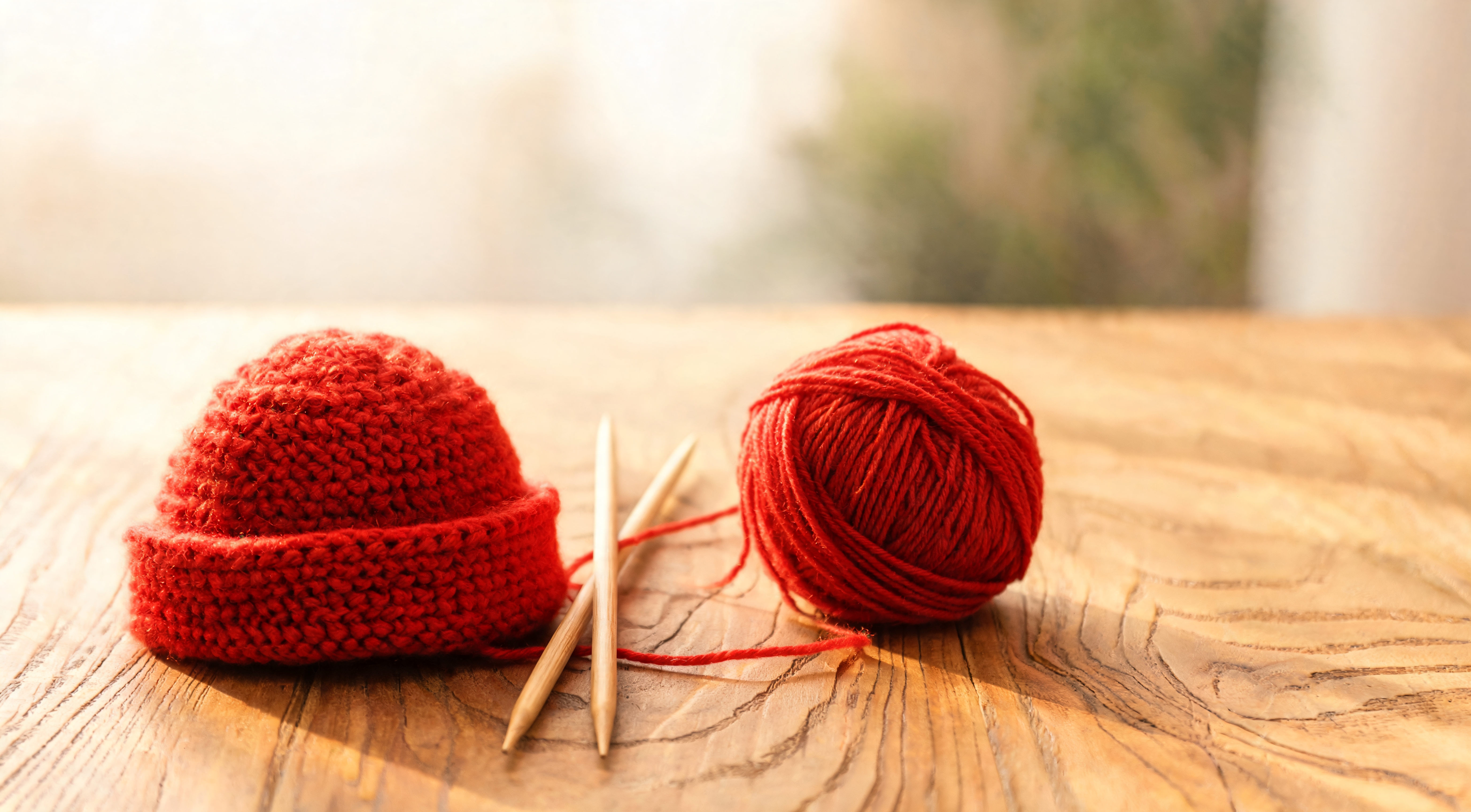 A small hand-knitted red roll-brim wool hat next to knitting needles and a ball of red yarn
