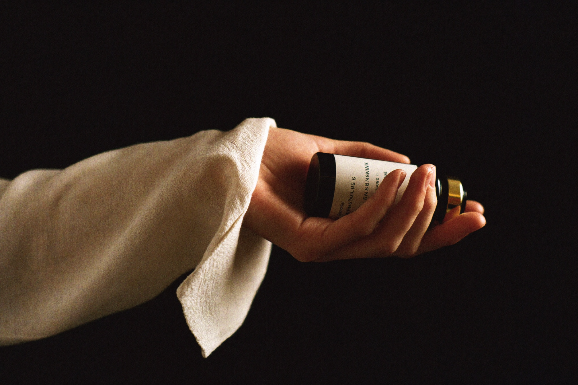 Intimate editorial photograph of a hand cradling a single dark-glass vial with a gold cap in warm low-key light.