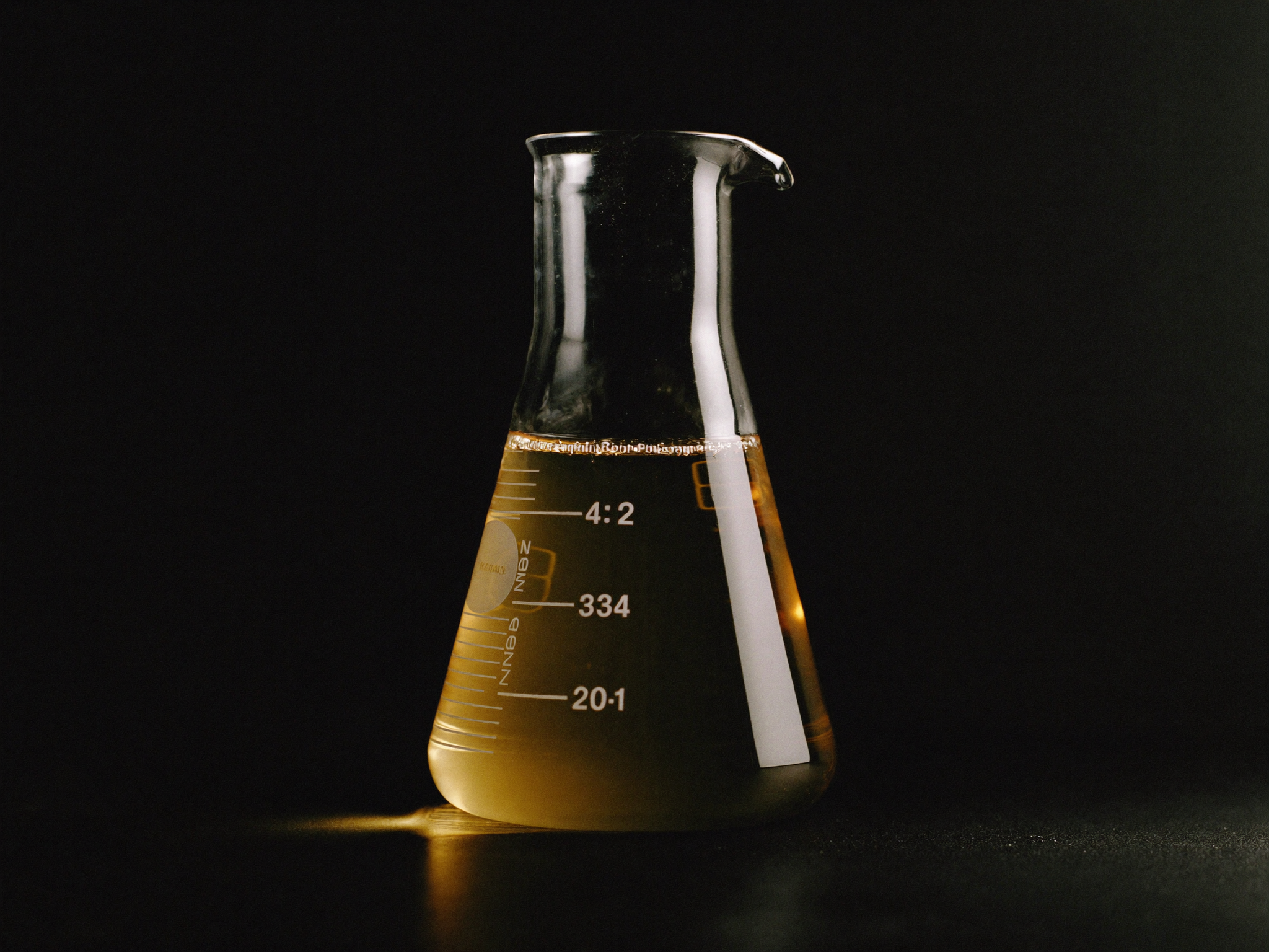 Editorial photograph of a scientific glass Erlenmeyer flask with amber-gold liquid on a matte black laboratory bench.
