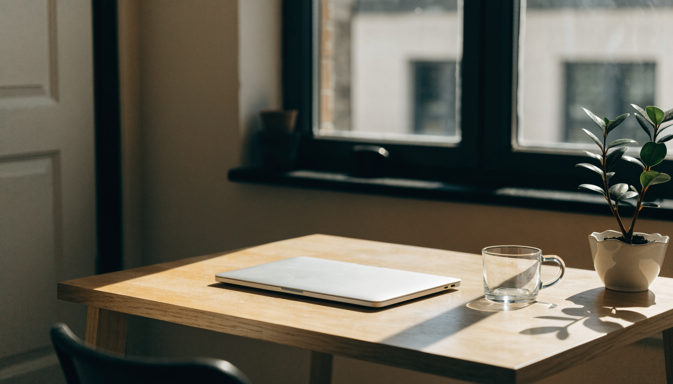 Bureau en bois clair près d une fenêtre avec un ordinateur portable fermé, une tasse vide et une plante verte