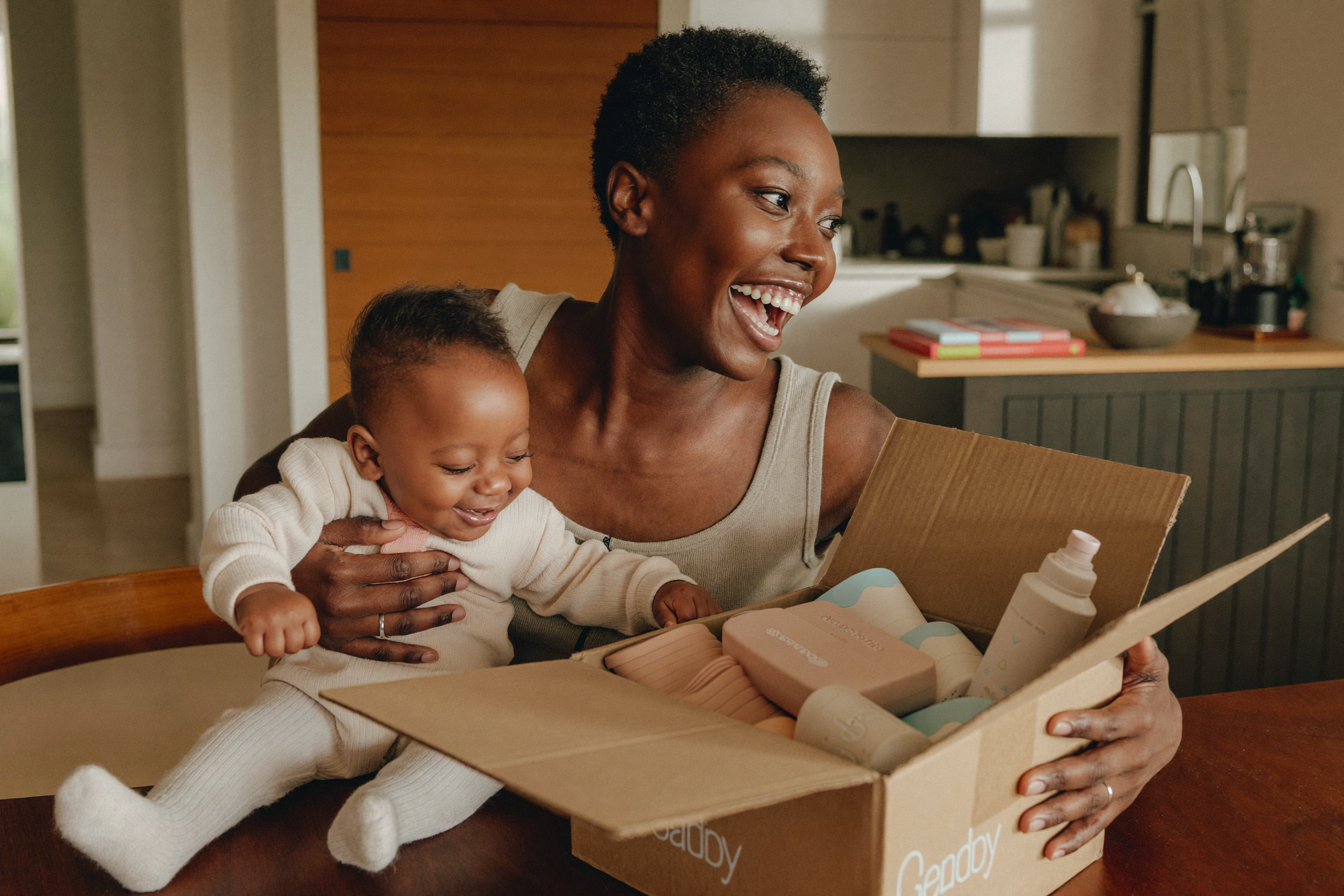 A smiling mum opening a box of baby products at the kitchen table with her baby on her hip
