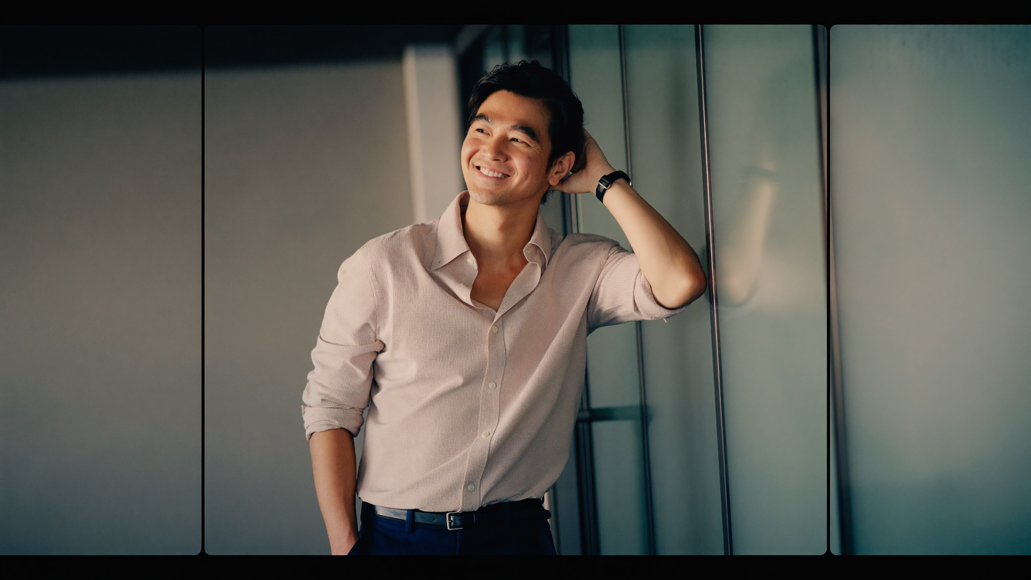 Chinese man in his 30s in smart casual attire leaning against a glass office wall, friendly relaxed smile