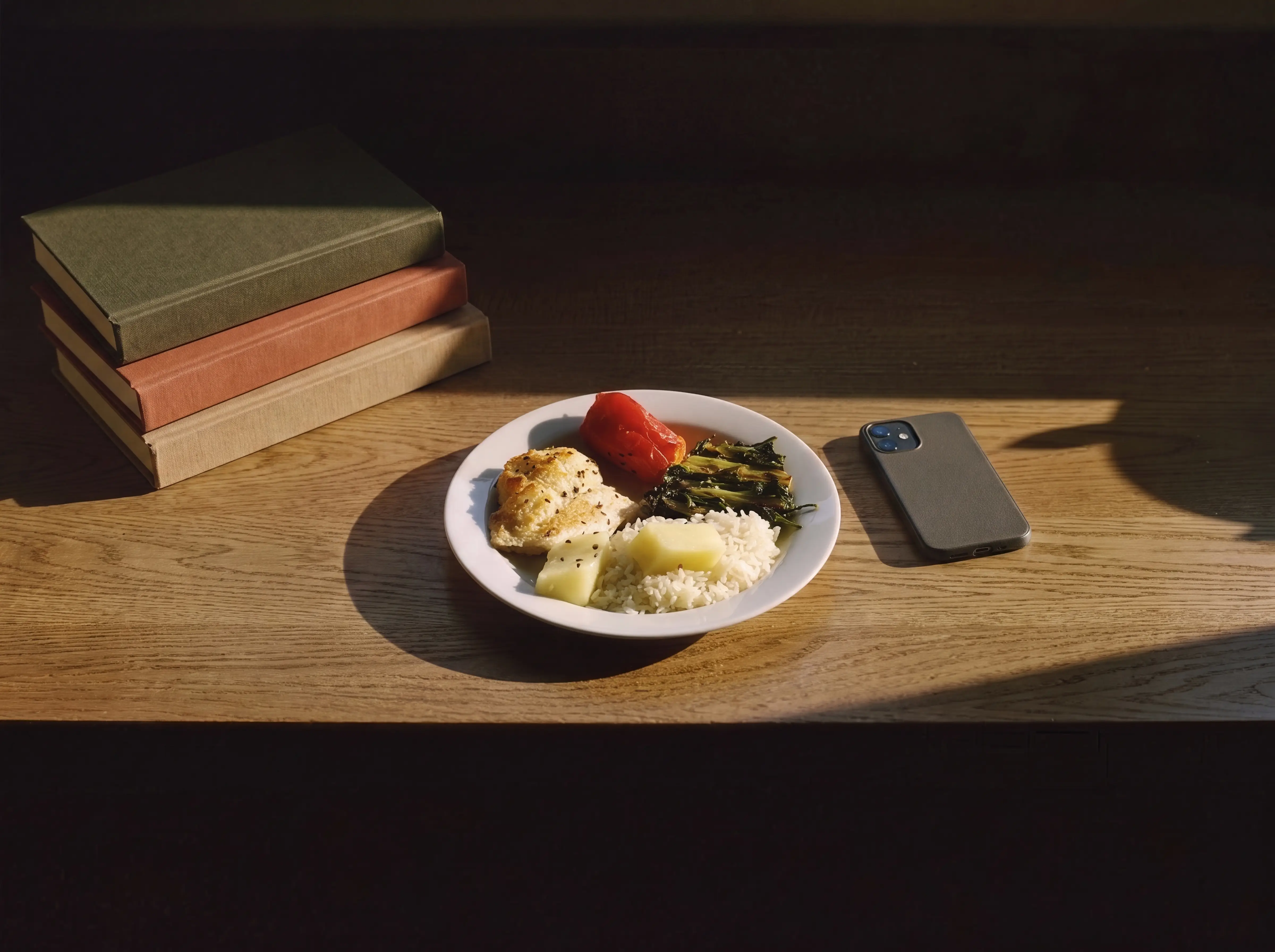 A plate of chicken, rice and vegetables resting alone on a desk beside a phone and a small stack of books
