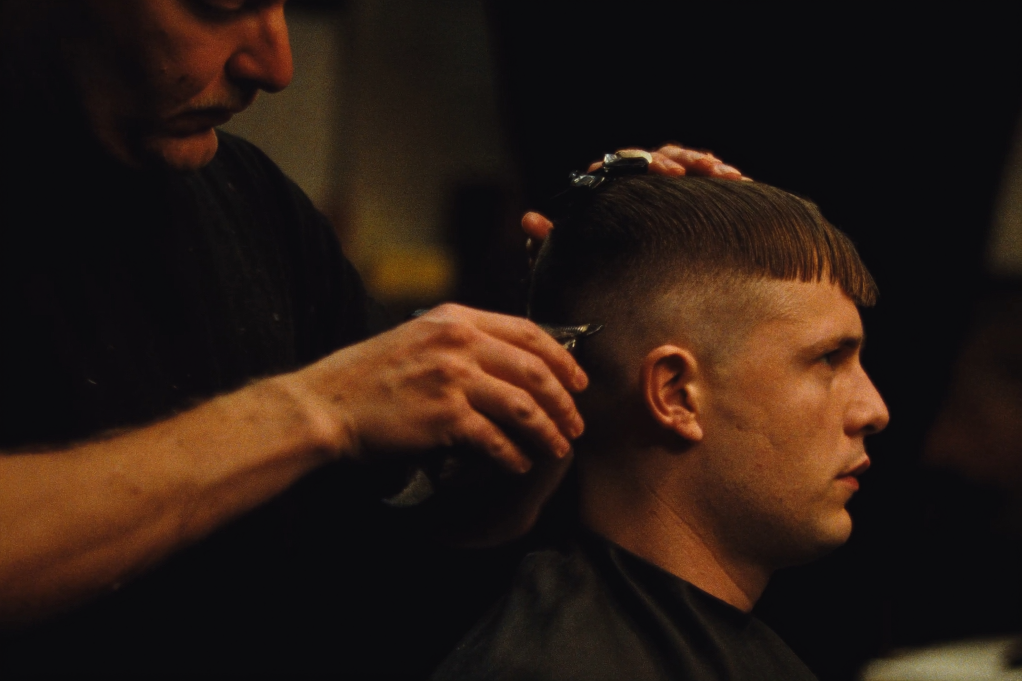 A barber blending a mid-fade with clippers under warm light