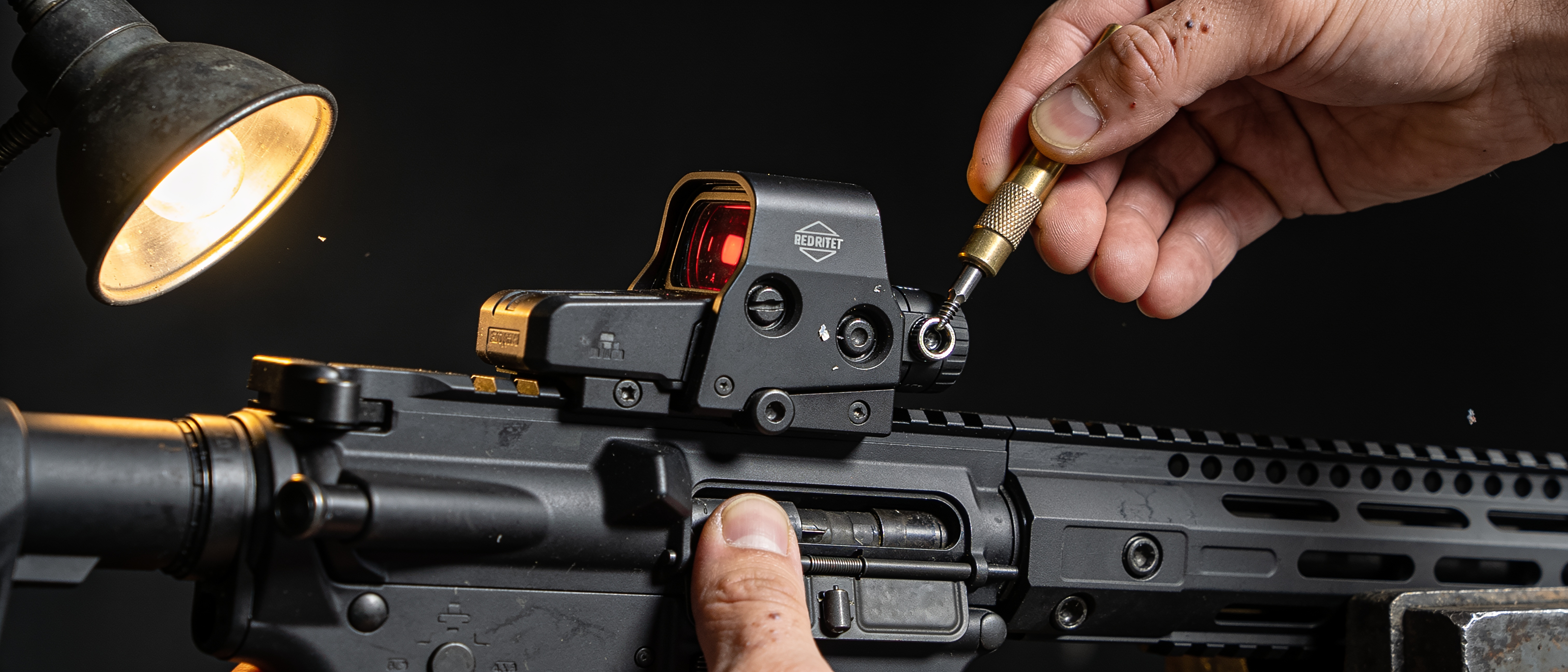 Hands torquing an optic mount onto a rifle receiver under a workbench lamp