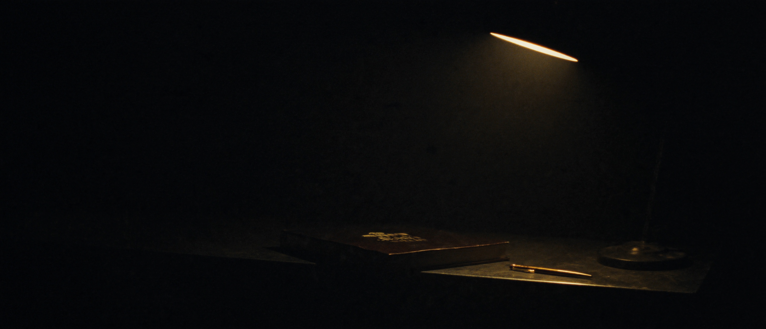 A bound record book and brass pen on a dark counter under a single task lamp
