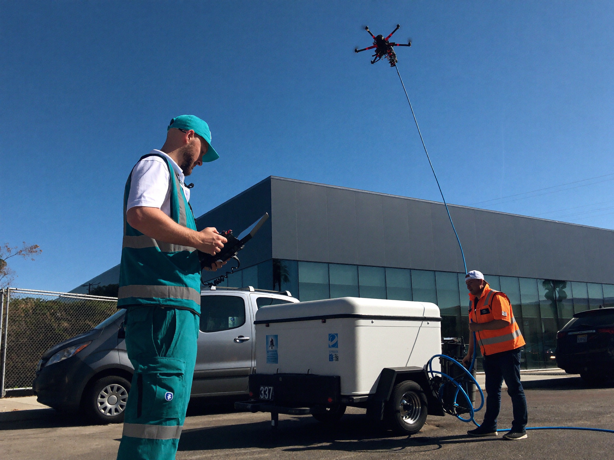 Humming Co. pilot and ground technician operating a cleaning drone from the sidewalk while it works a glass facade above