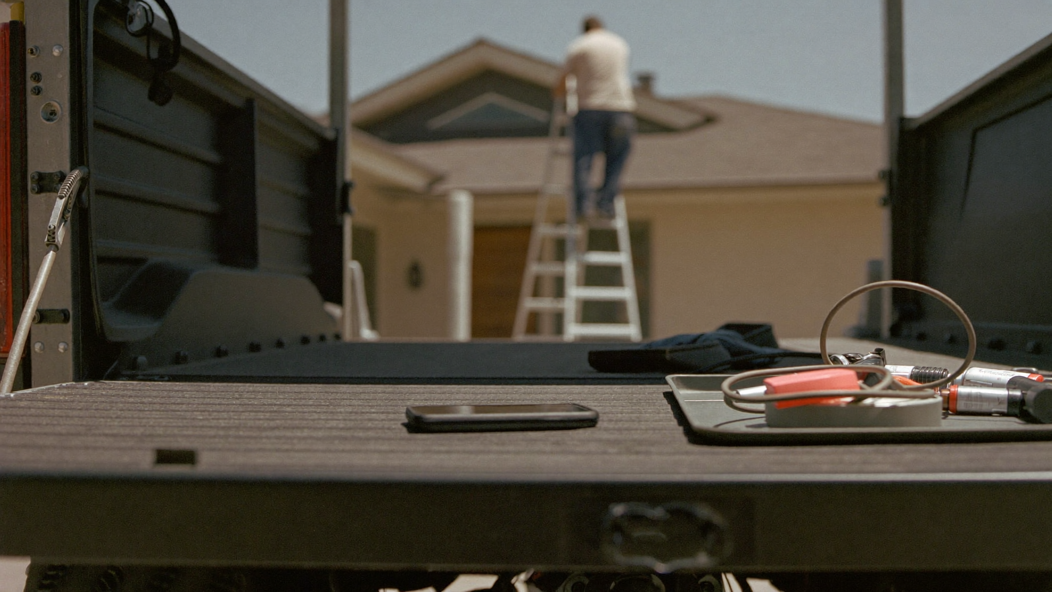 A phone left face down on a work ute tailgate among tools while a tradesperson works up a ladder in the background