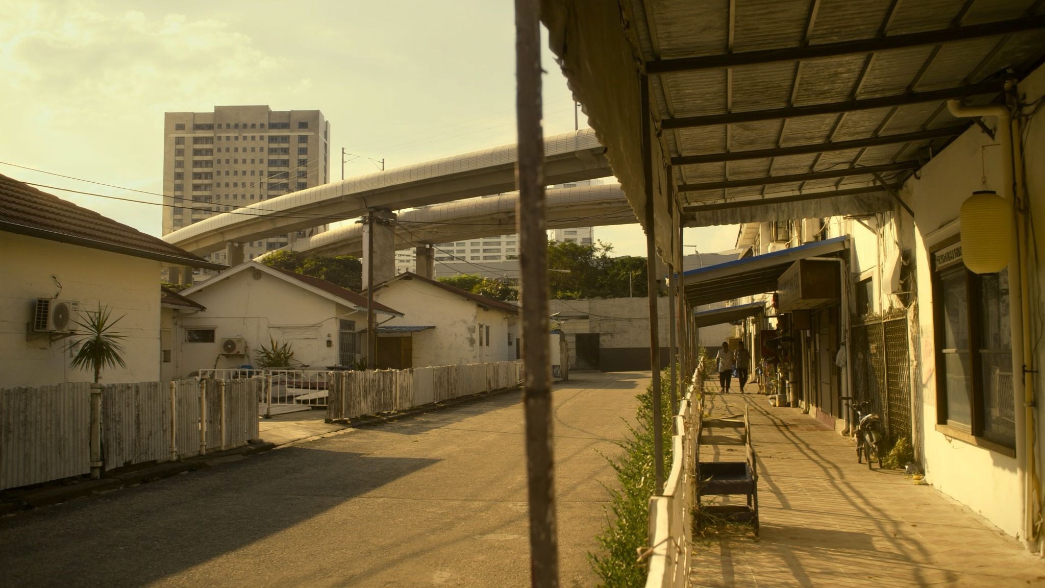 A covered shophouse footpath in Johor Bahru leading toward an elevated transit rail line in golden afternoon light
