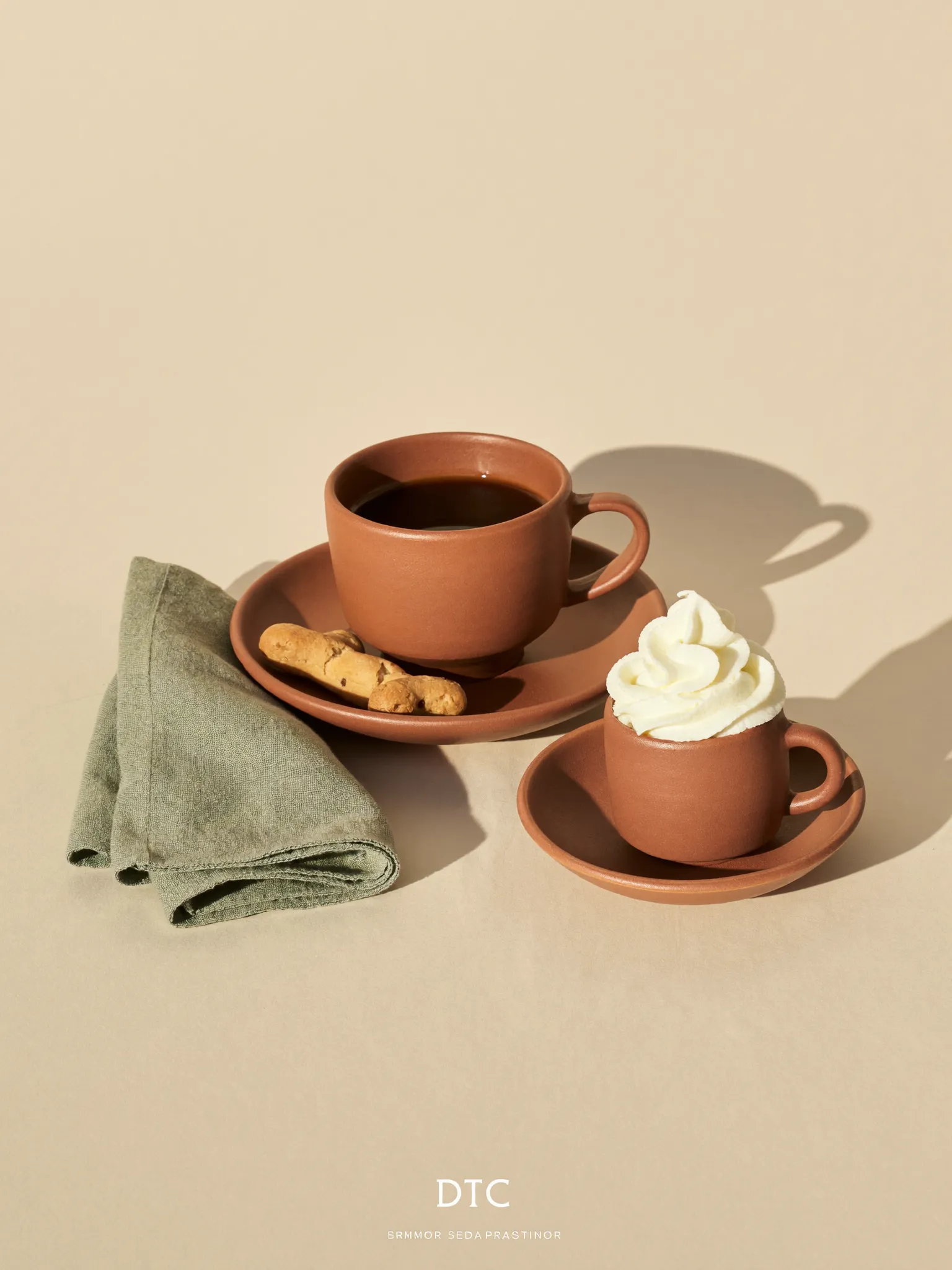 A terracotta cup of long black coffee beside a tiny puppuccino with a bone-shaped dog biscuit, on a warm cream background