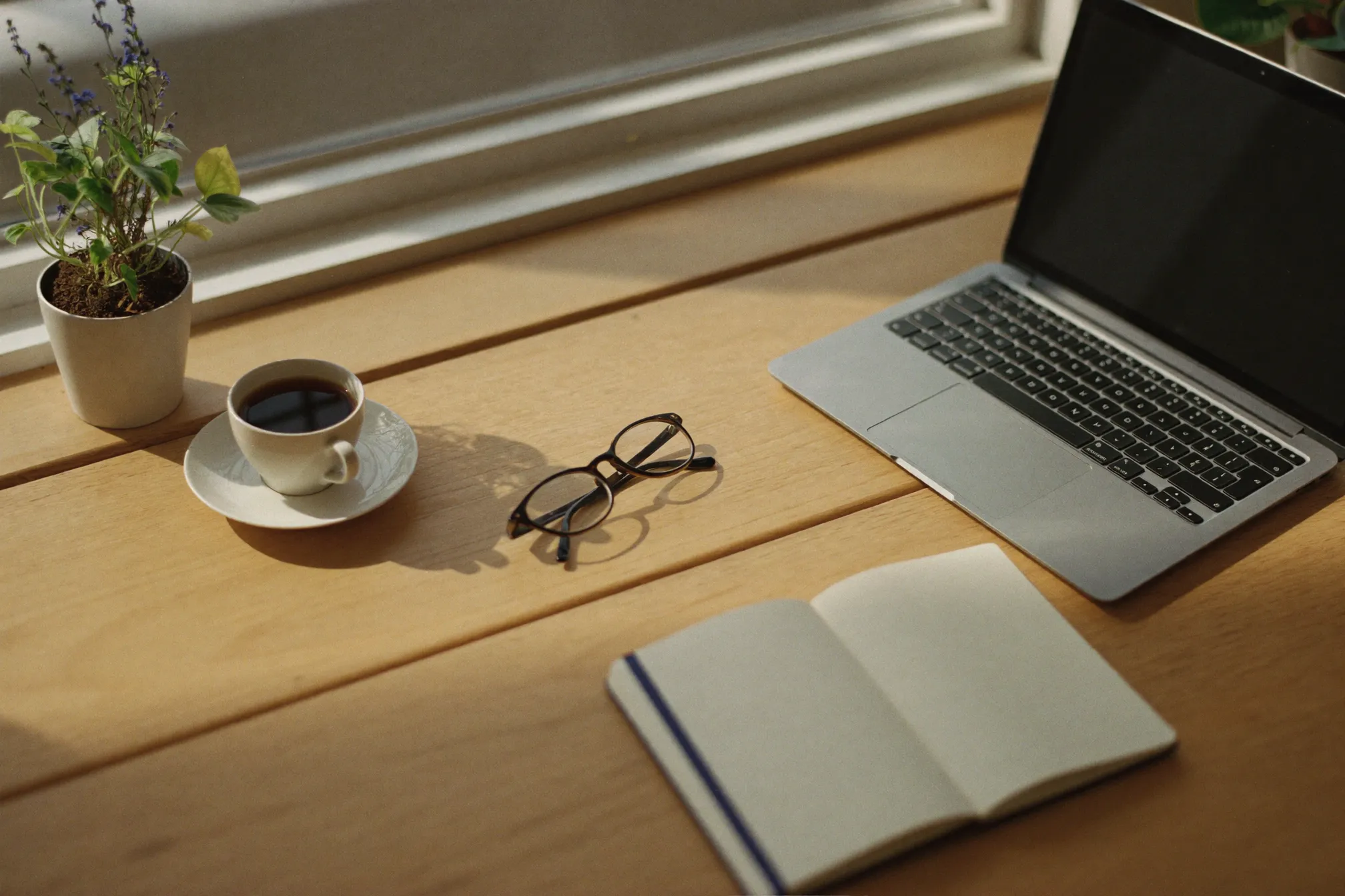 Overhead still life of a closed laptop, coffee cup, plant and notebook on a pale oak counter