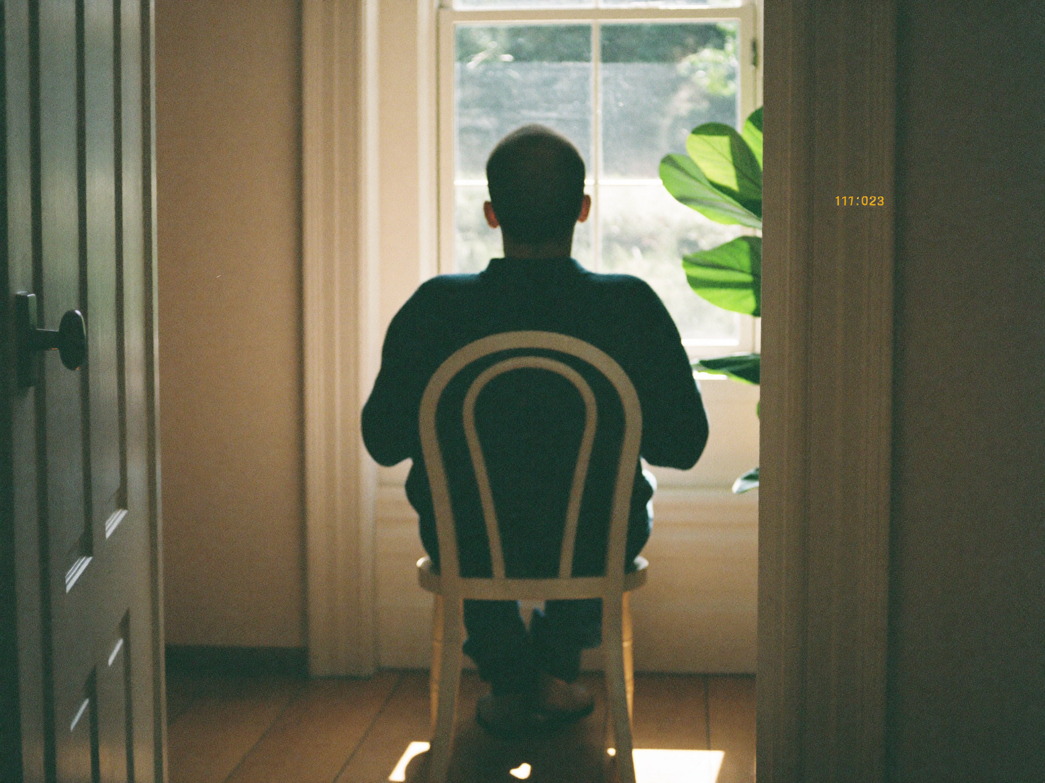 A person at rest in a chair by a tall window, seen from behind