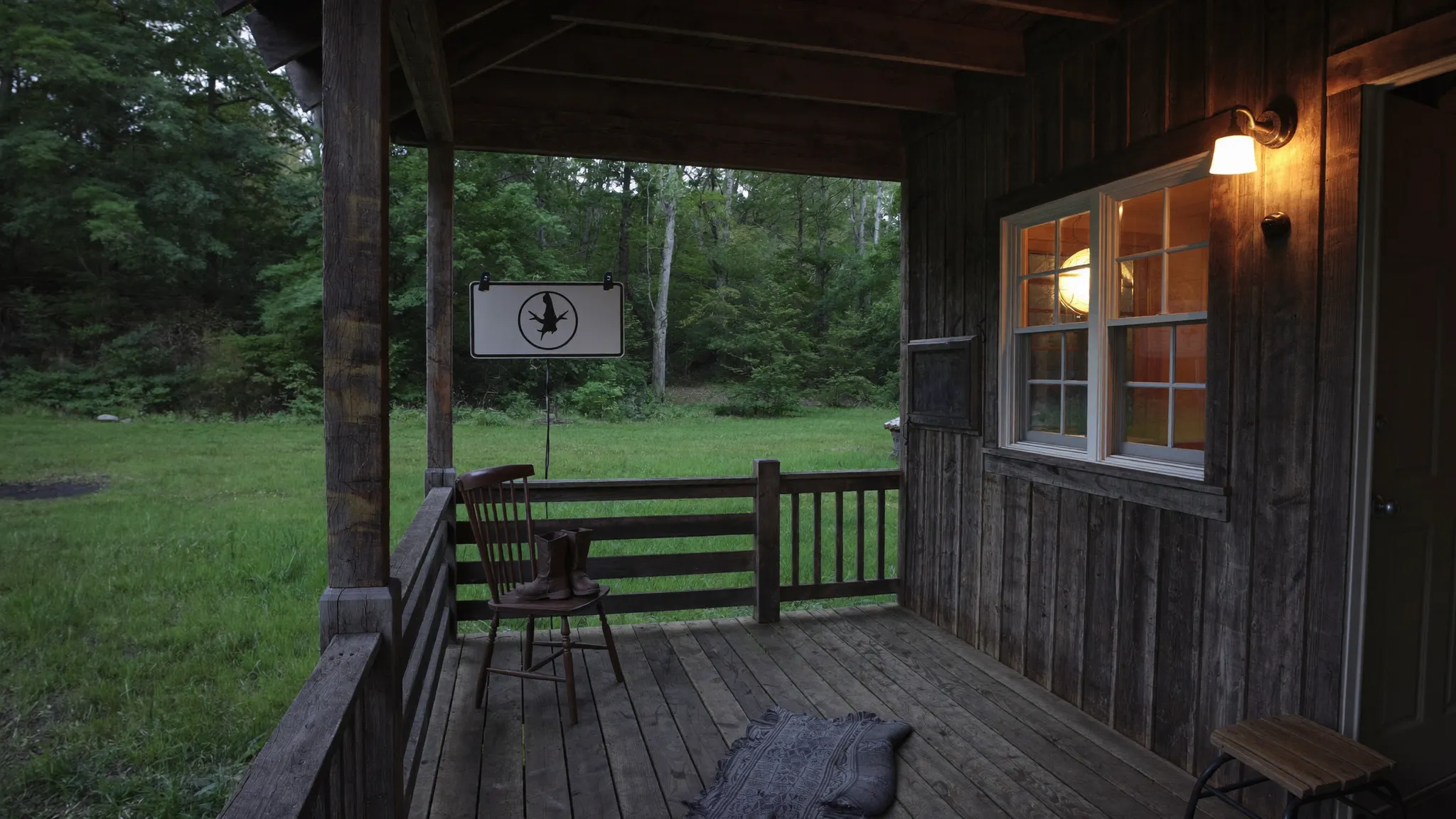 A weathered hunting cabin porch at dusk with worn boots on a chair and lamp light in the window