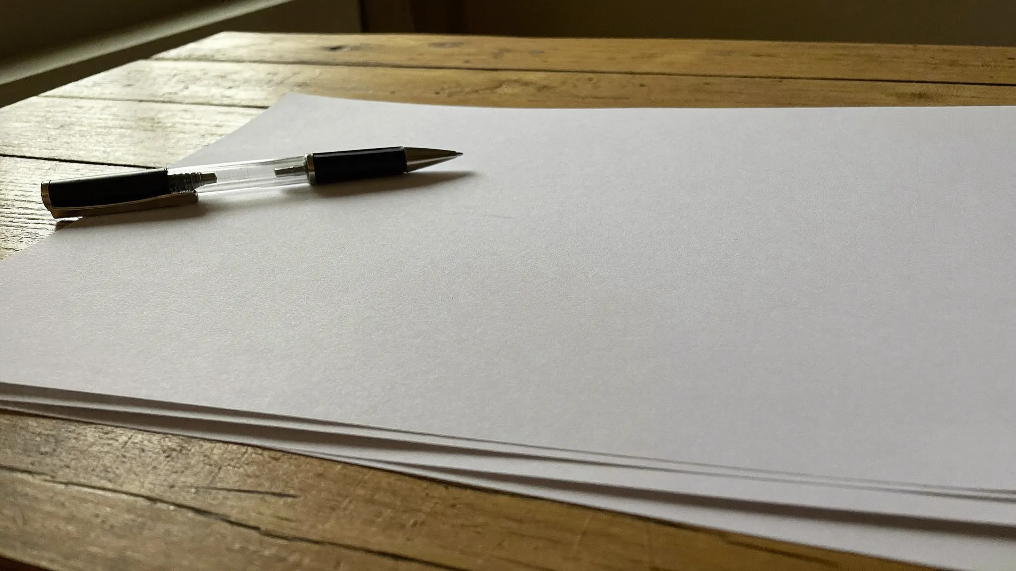 A pen resting on a stack of blank paper on a wooden farmhouse table in warm window light