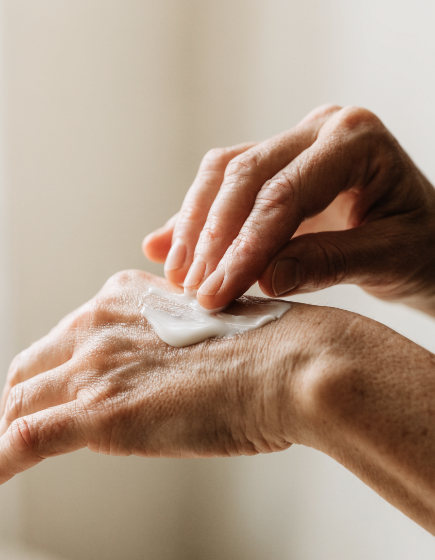 Two mature hands, one smoothing cream across the back of the other