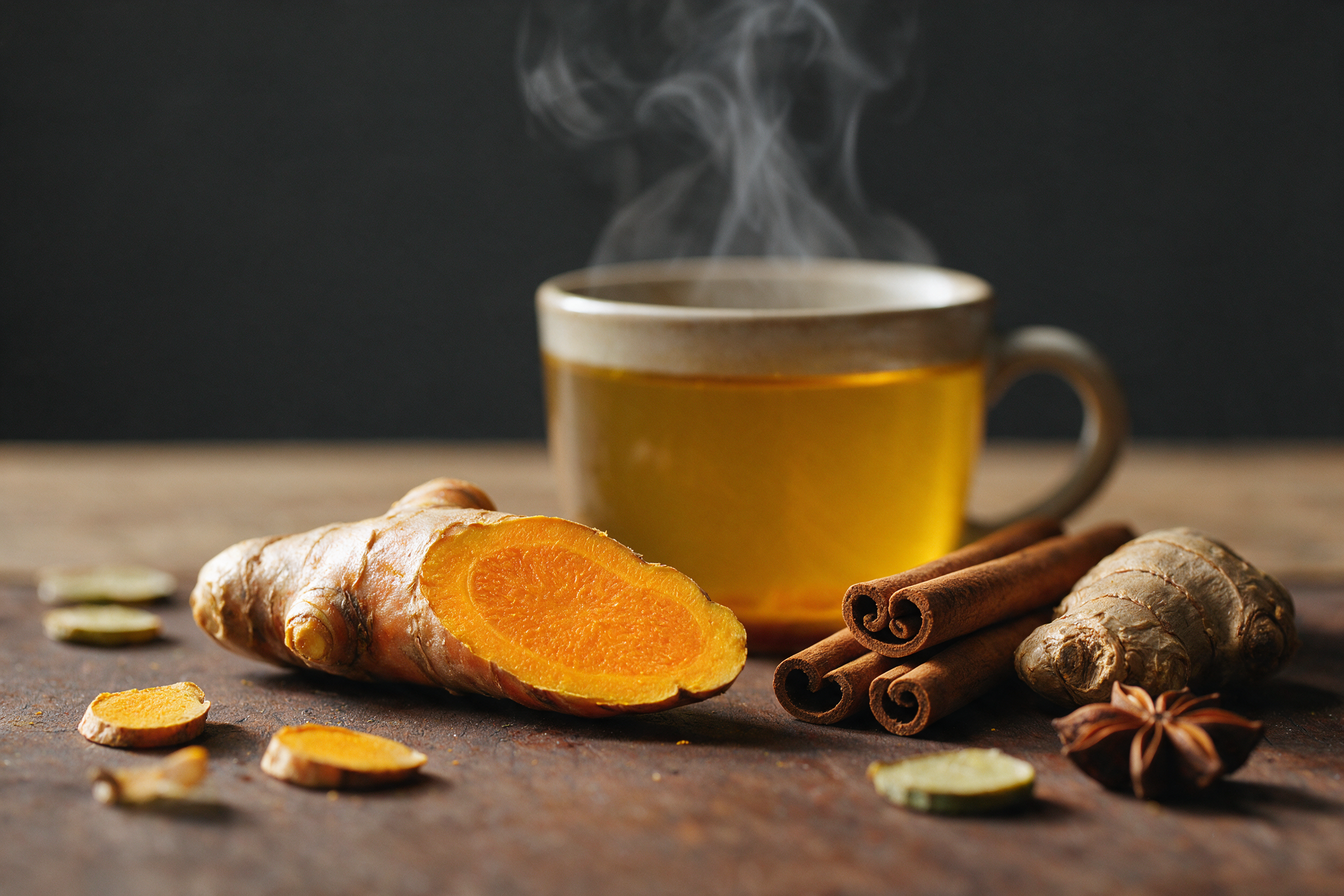 A sliced turmeric root revealing its vivid orange interior, next to cinnamon quills, ginger and a steaming cup of tea