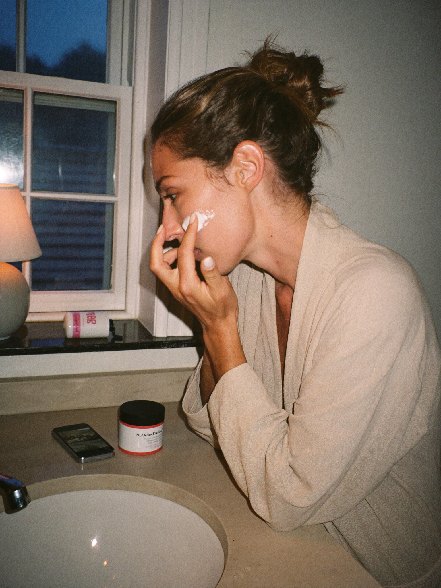 A woman at a stone bathroom sink in the evening, pressing night cream into her cheek, her phone face-down on the counter.