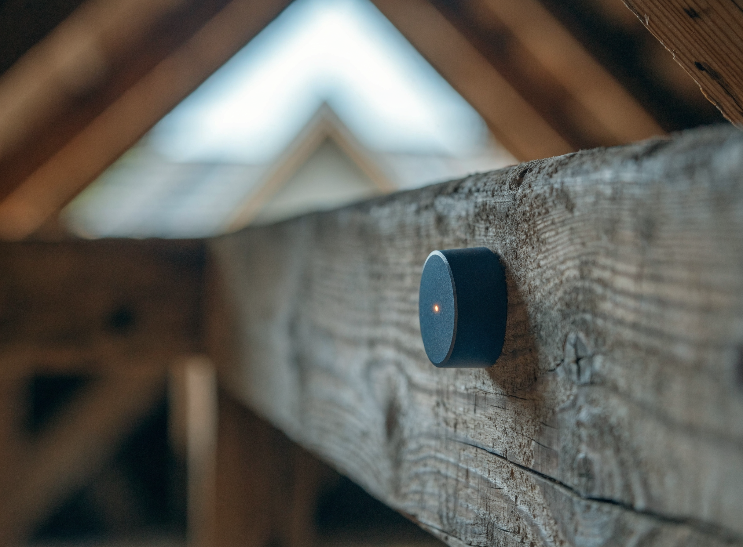 Small wireless navy Attic Vitals sensor mounted on a wooden attic rafter with a soft amber status light