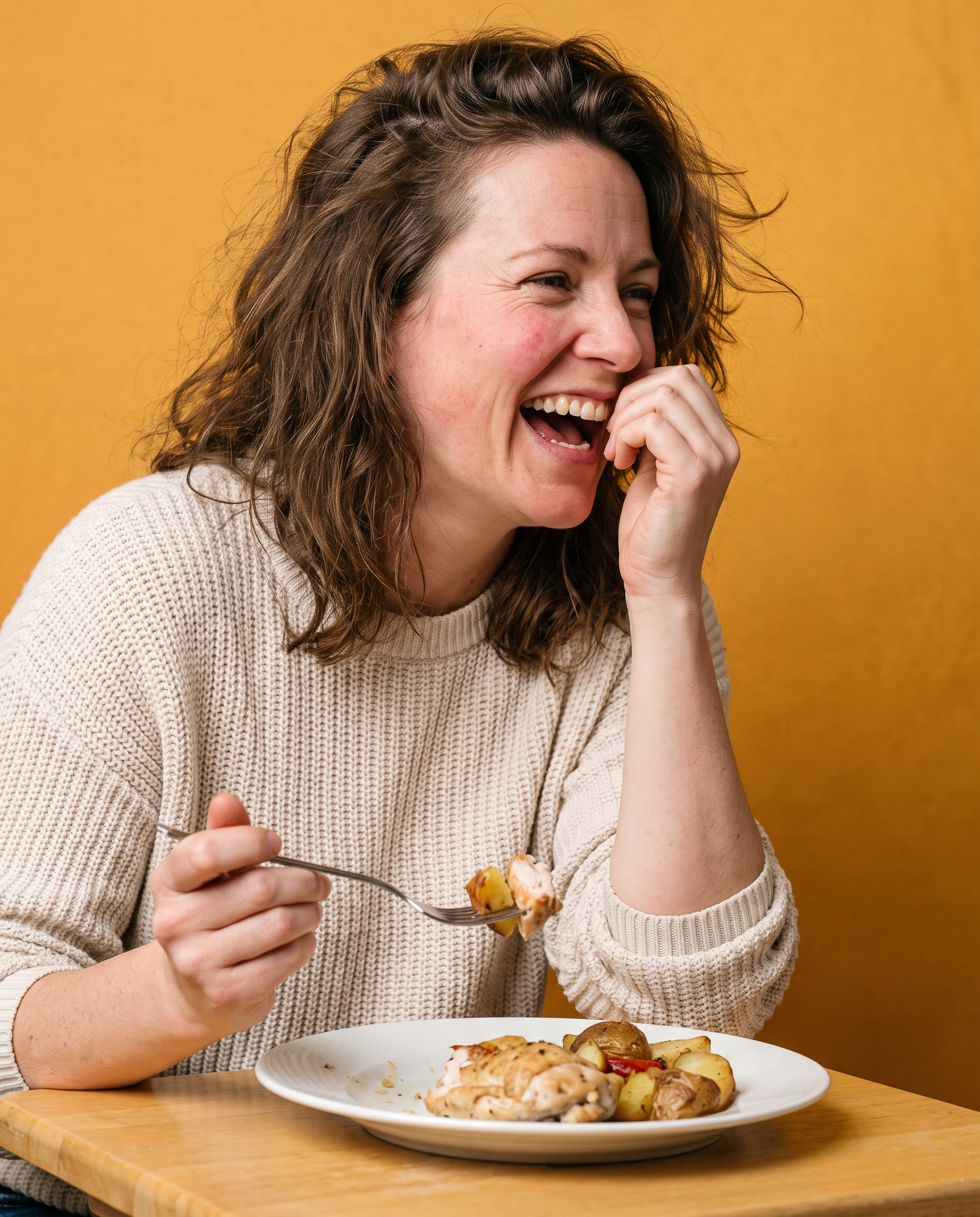 A woman mid-laugh at a table against amber seamless, fork halfway down