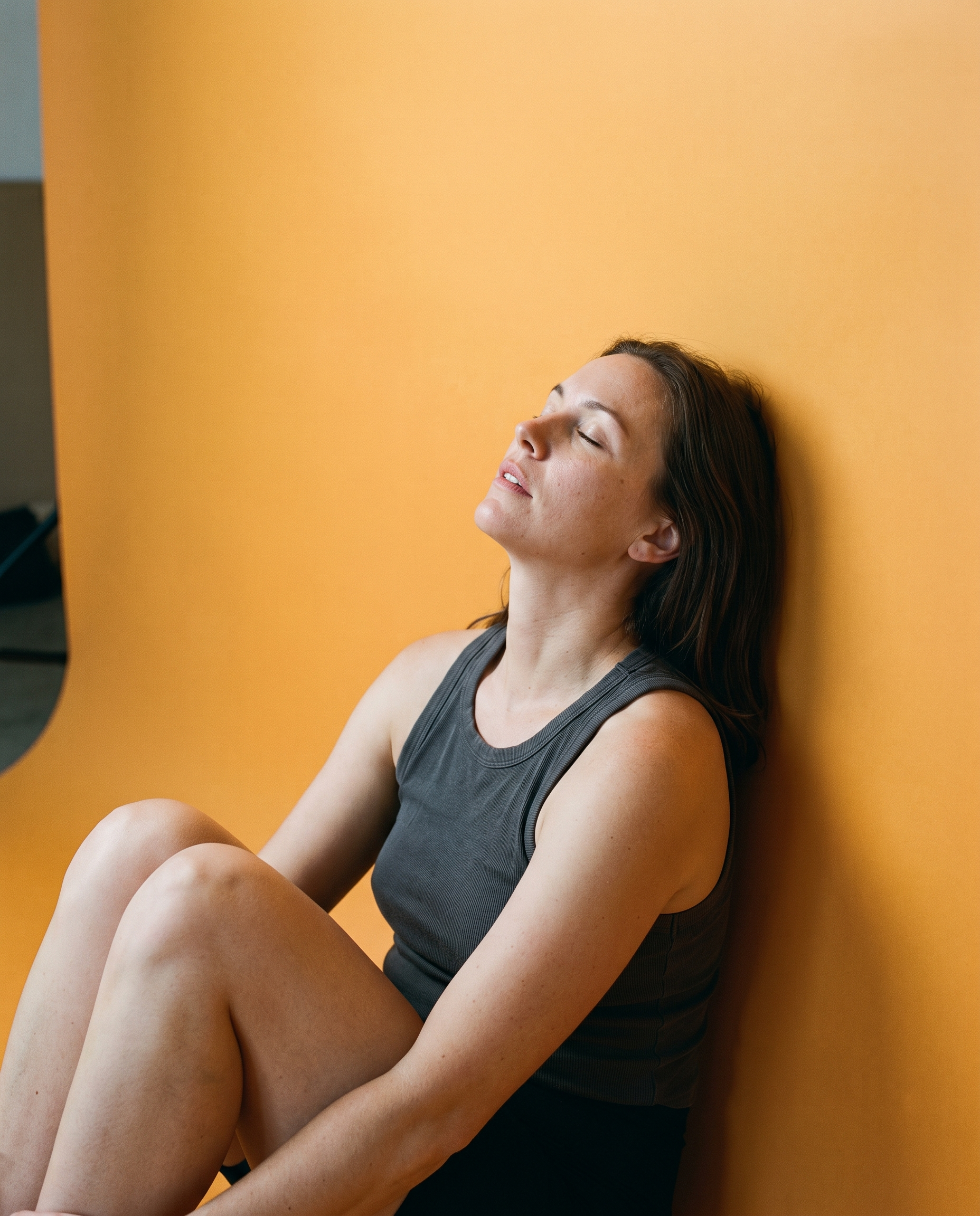 A woman sitting at rest against an amber seamless cove, eyes closed