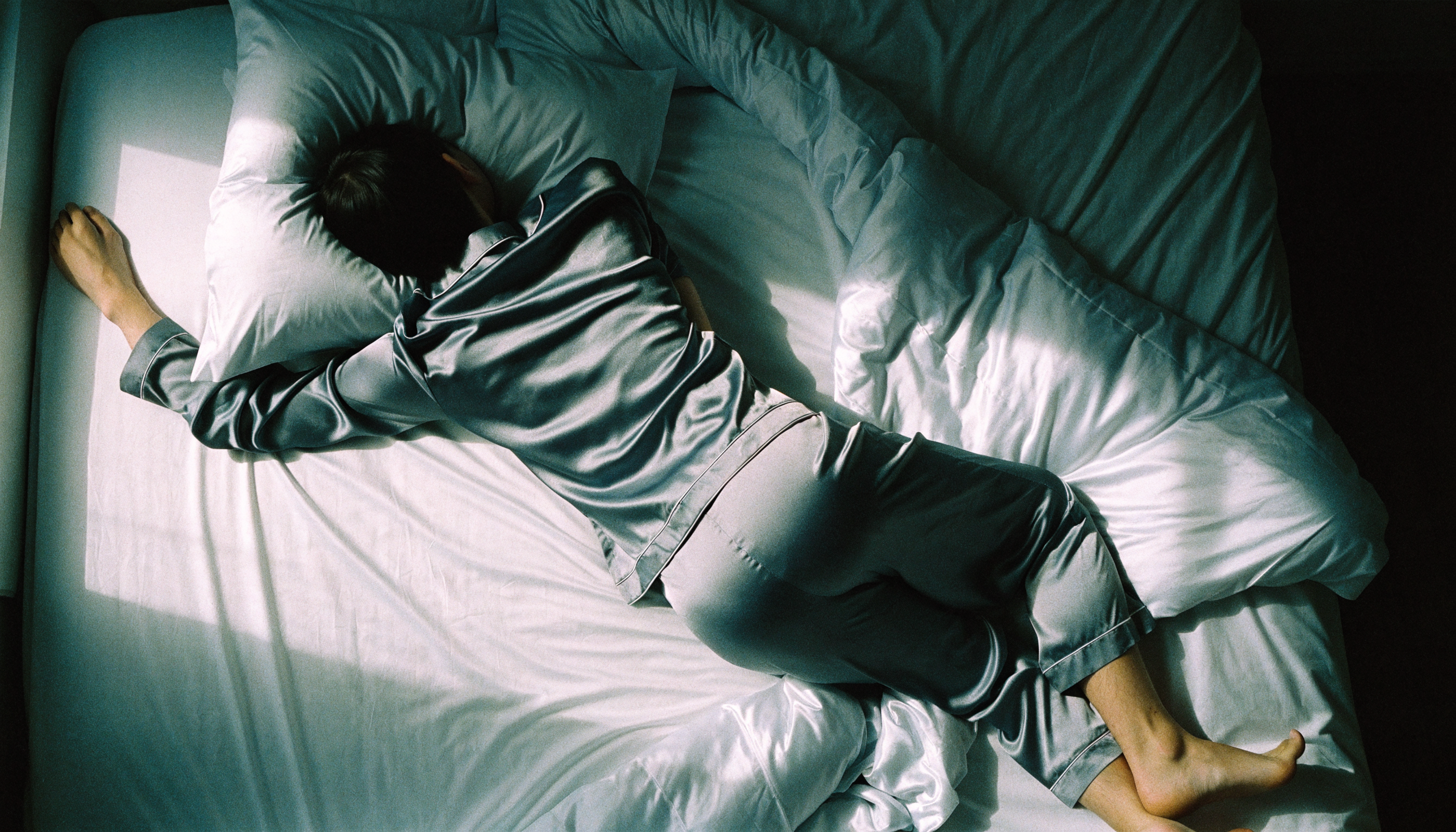 A restless sleeper at first light in synthetic satin pyjamas, duvet kicked down